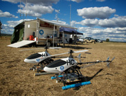 Unmanned helicopters in a field with vehicles and shelters in the background.