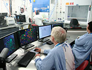Scientists sitting in front of a bank of computers watching other scientists work in a secure laboratory environment.