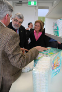 Looking over the shoulder of a man resting his hand on a box of nappies, two people listen.