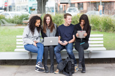 Group of young adults sitting at bus stop using a range of electronic devices.