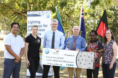 Six people standing in a treed garden, holding an oversized cheque in front of flags and CSIRO information panel poster.