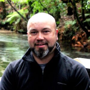 Head and shoulders photo of Bradley Moggridge standing in nature with a stream and vegetated bank in the background. 