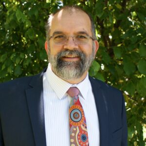 Head and shoulders photo of Peter Radoll standing in front of shrubs. 